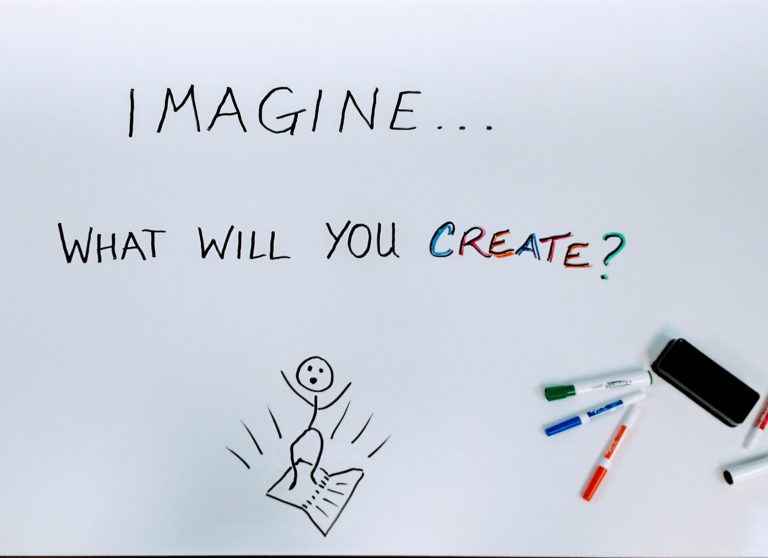 A whiteboard tabletop with the handwritten words "Imagine... what will you create?". Below the words, a stick is jumping in surprise out of a book. Dry erase markers and an eraser are scattered in the corner.