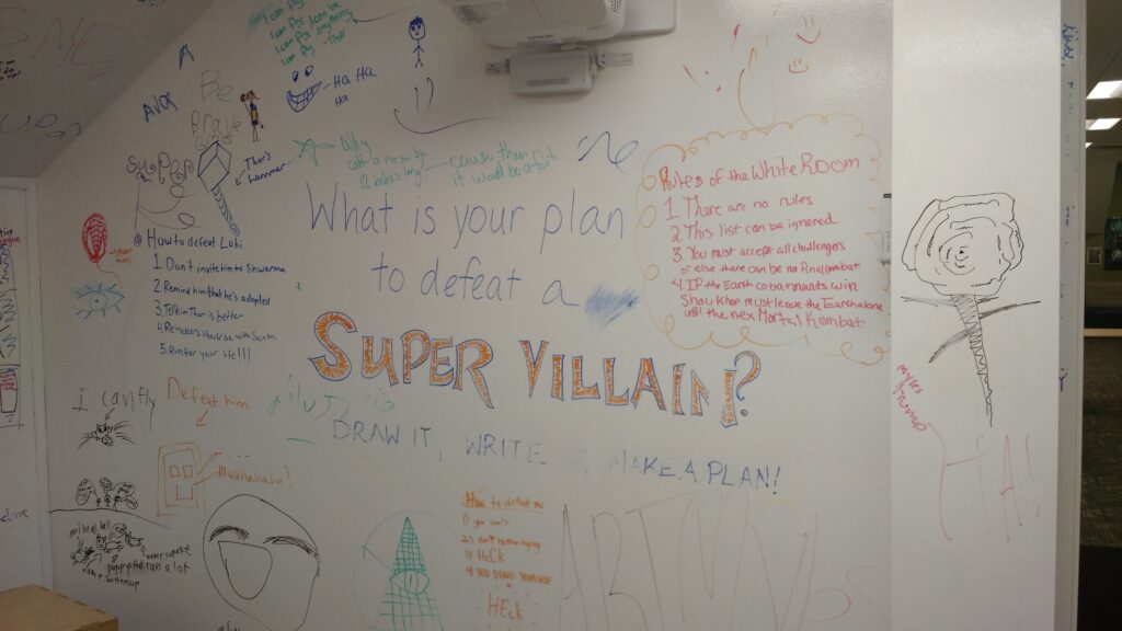 A whiteboard wall. Text in the middle reads "What is your plan to defeat a super villain? Draw it. Write it. Make a Plan." Around those words the wall is filled with drawings and written plans created by many different people.